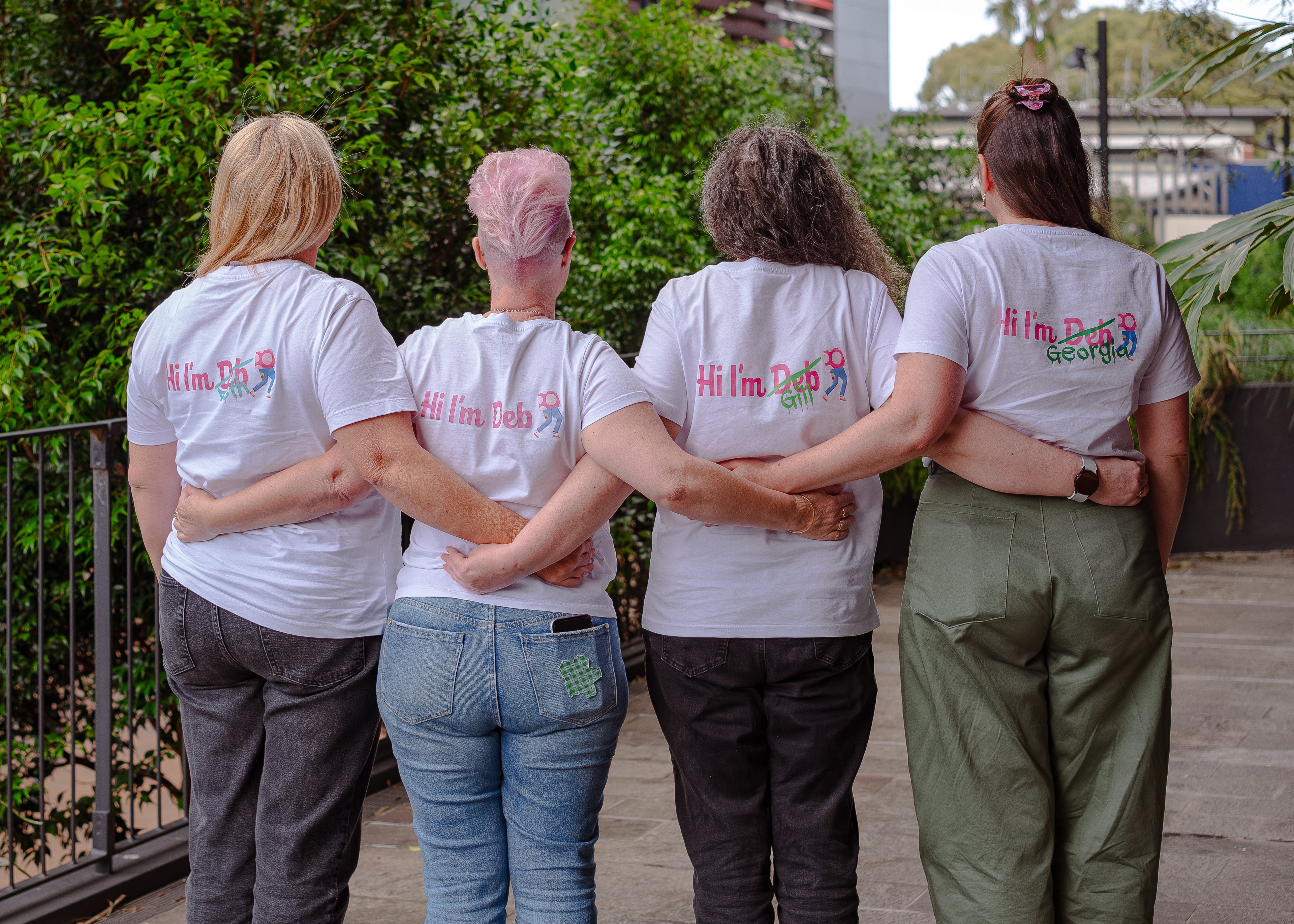 Four women standing with their arms around each other, wearing matching white t-shirts with colorful text.