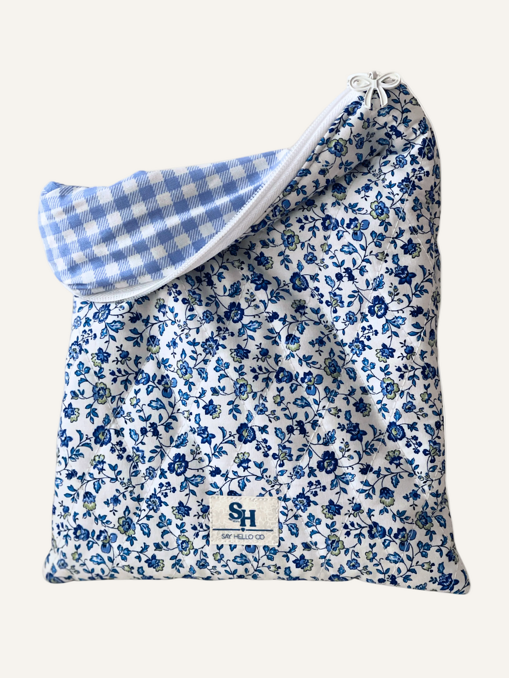 a quilted book sleeve in ditsy blue floral on white fabric with blue gingham lining and a white bow zipper on a warm cream background