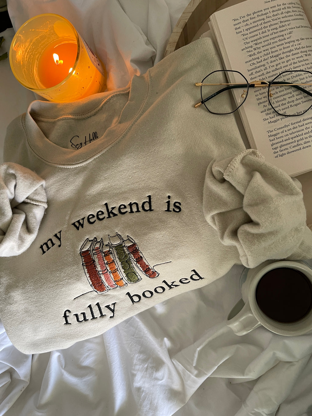 A cozy beige crewneck sweater with the phrase 'my weekend is fully booked' embroidered on the front, displayed alongside a cup of coffee and a book, suggesting a relaxed, leisurely weekend atmosphere.