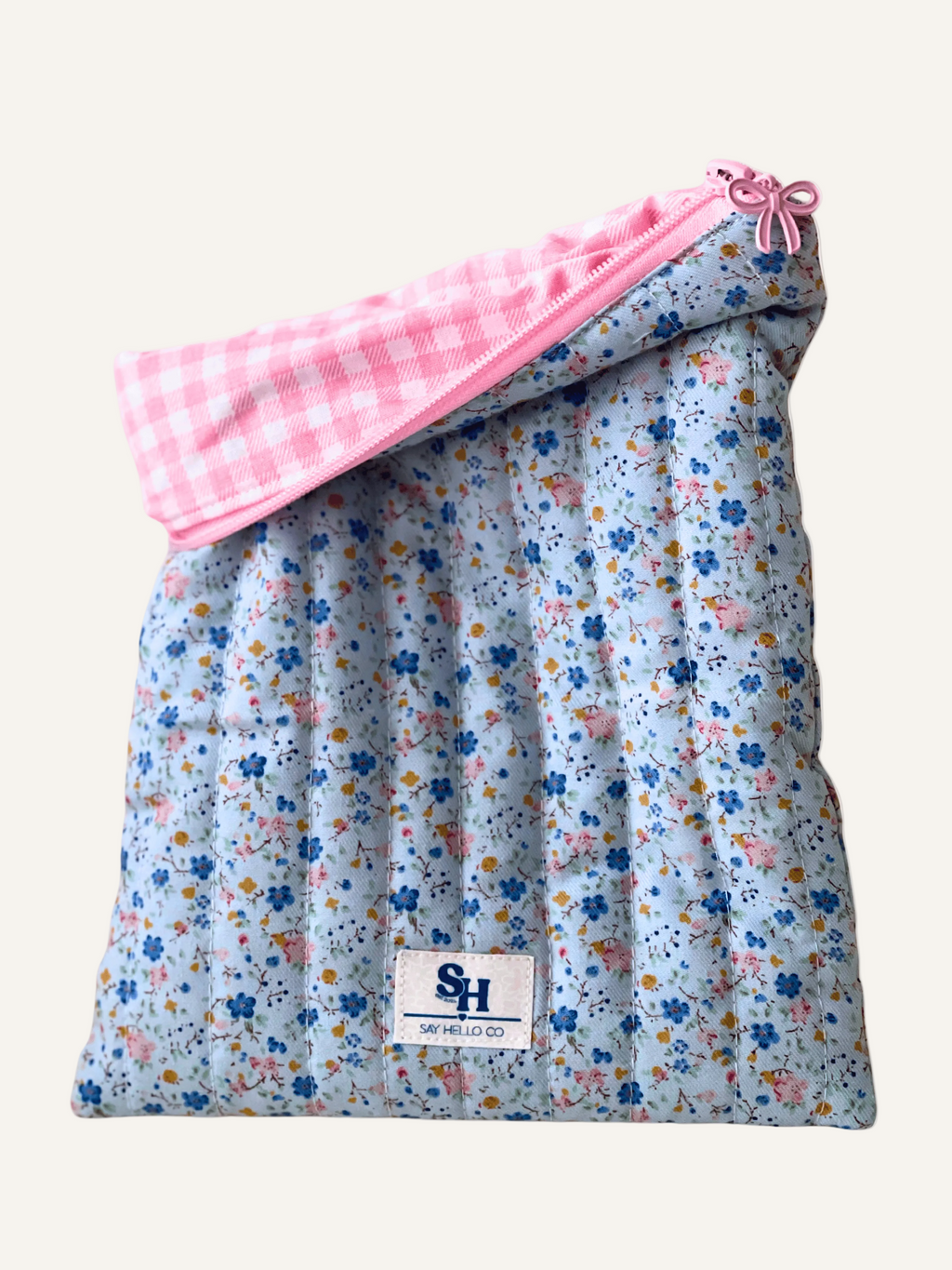 a quilted book sleeve in soft light blue with ditsy blue and pink florals fabric with a pink bow zipper and a pink gingham lining on a warm cream background