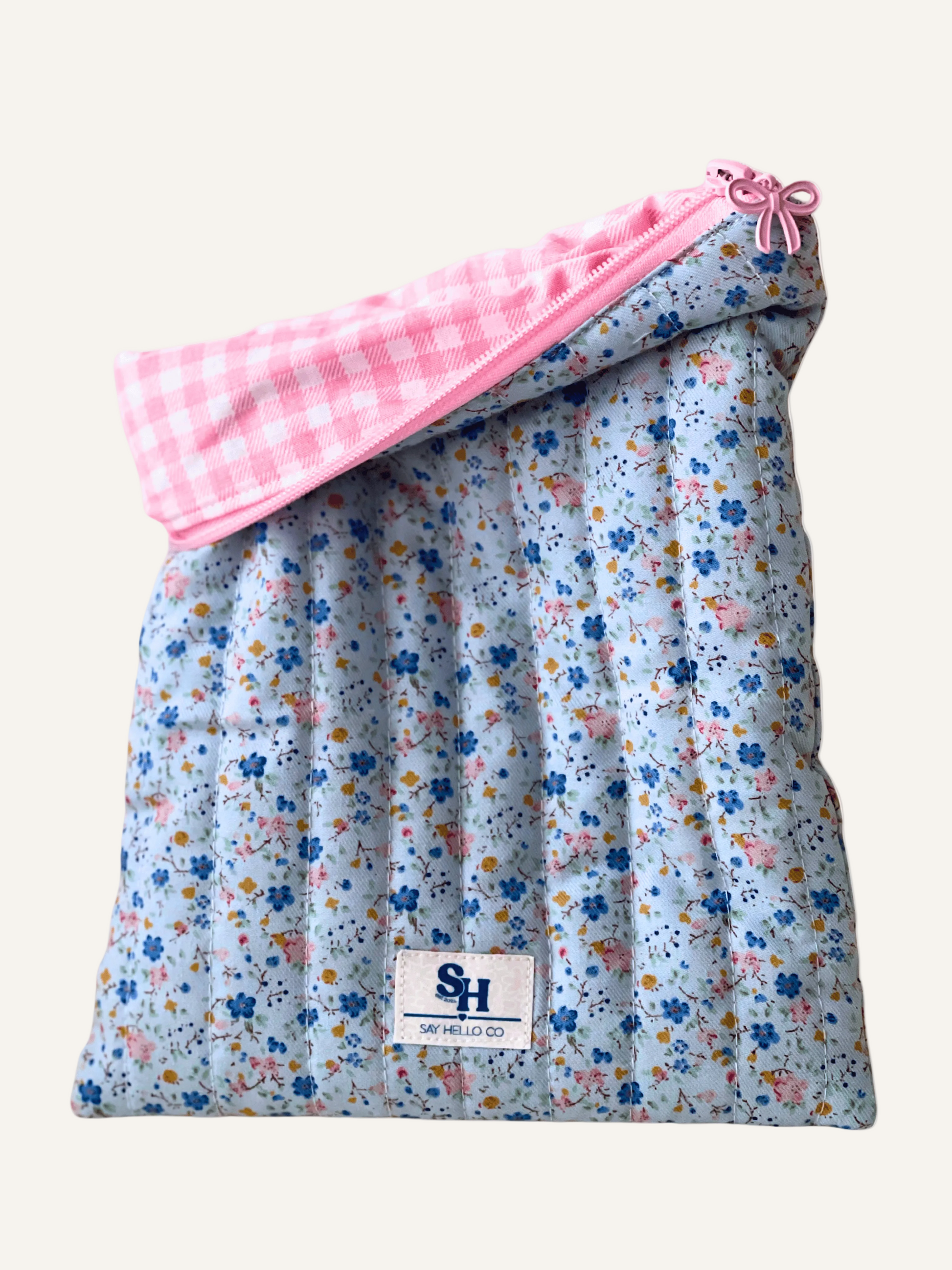 a quilted book sleeve in soft light blue with ditsy blue and pink florals fabric with a pink bow zipper and a pink gingham lining on a warm cream background