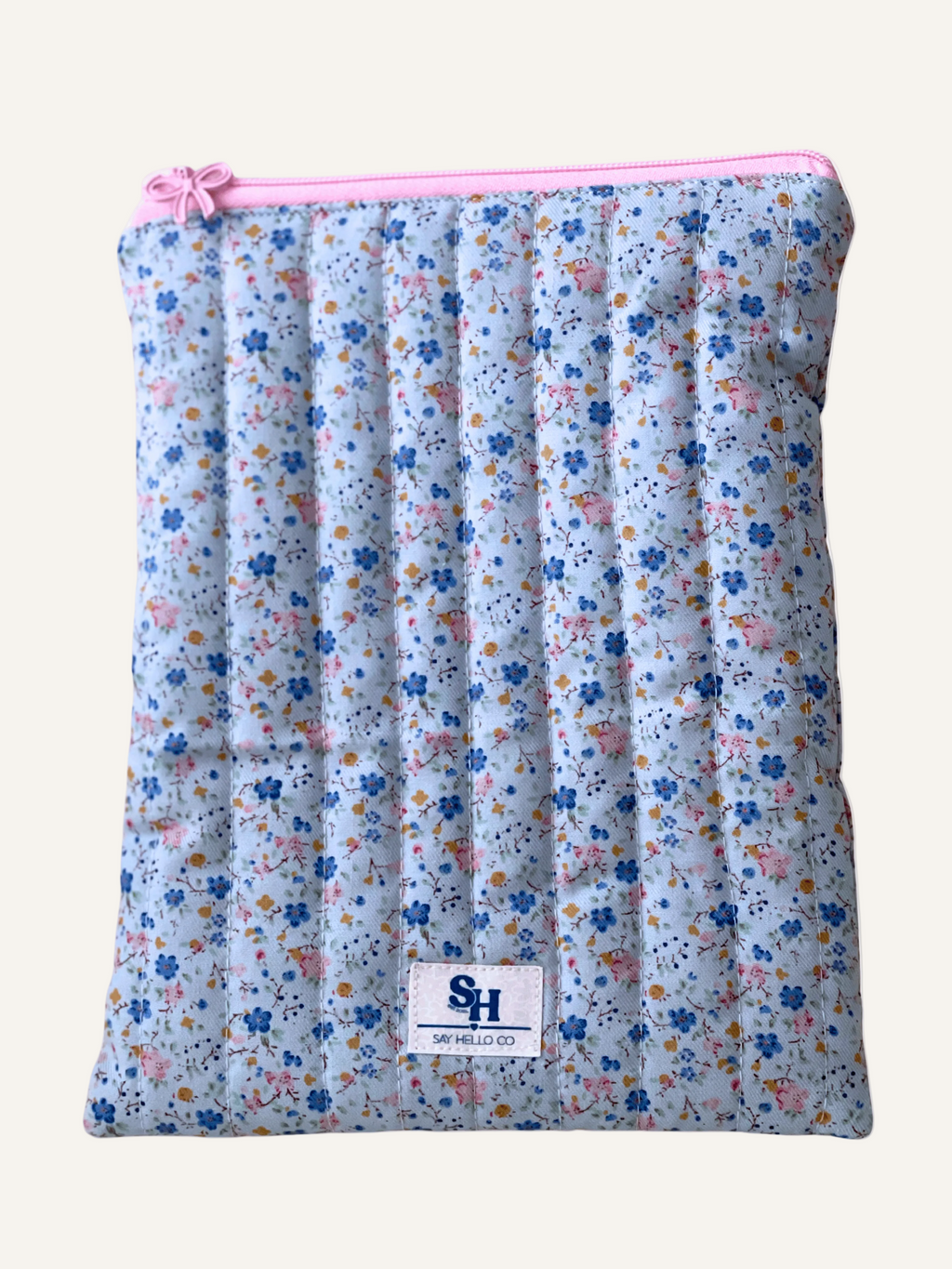a quilted book sleeve in soft light blue with ditsy blue and pink florals fabric with a pink bow zipper on a warm cream background