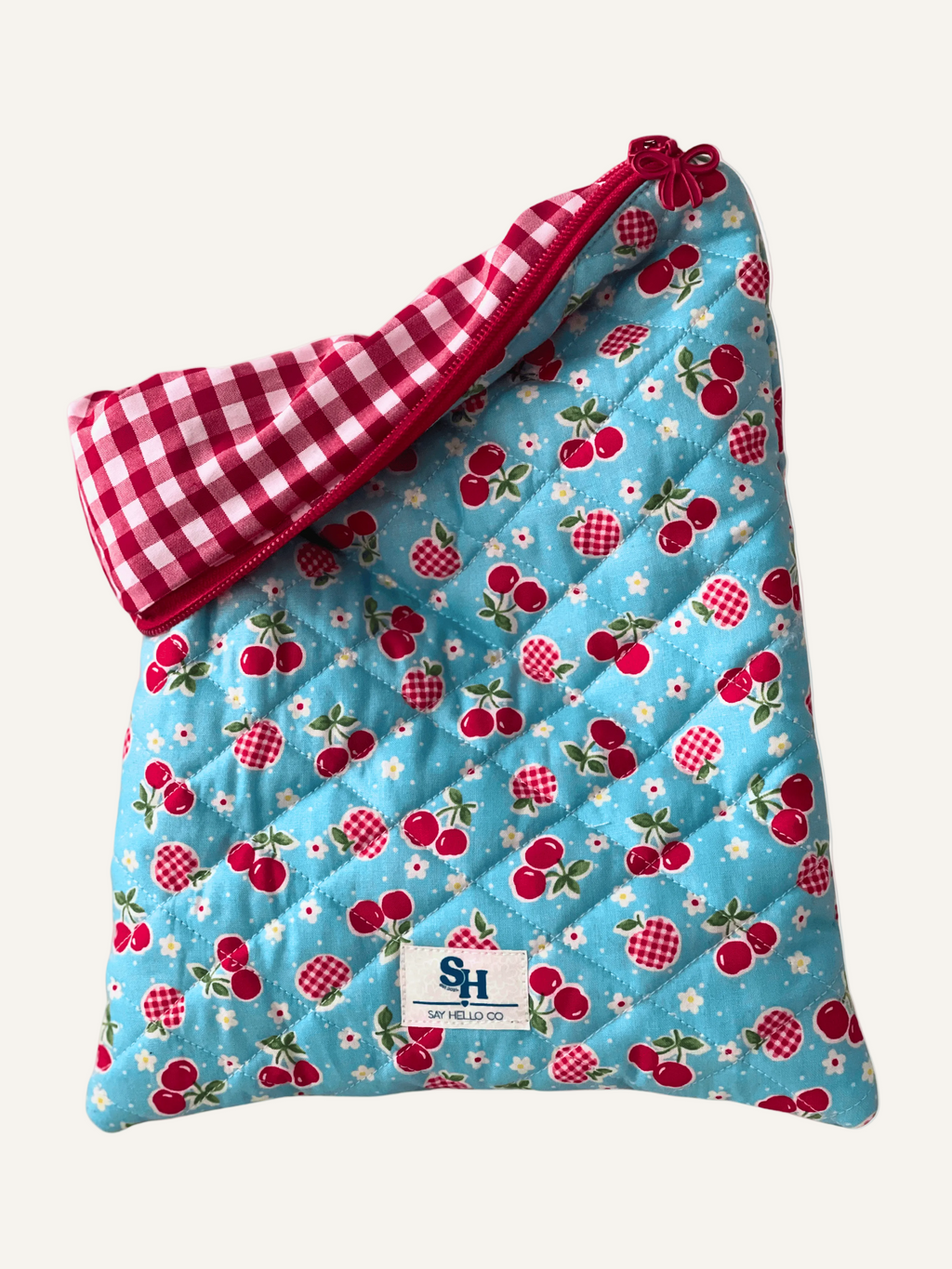 Quilted book sleeve with cherry and gingham apple pattern on sky blue fabric and red gingham lining on a warm cream background