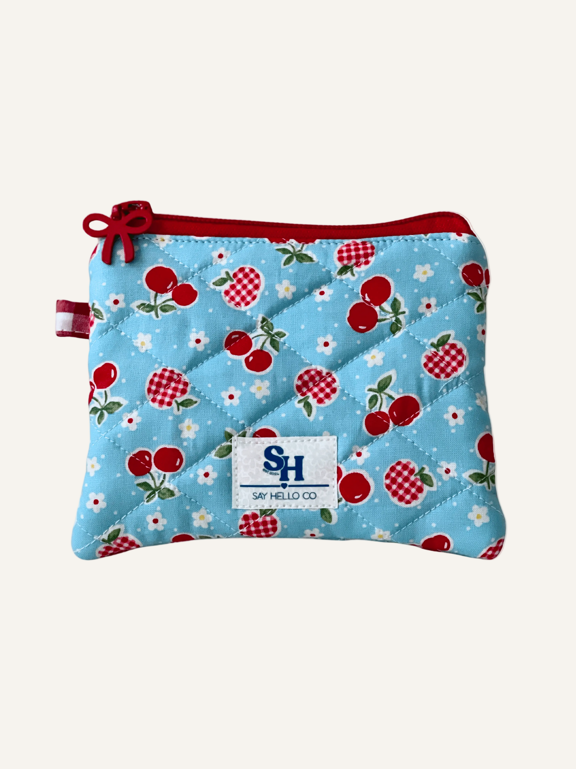 Small light blue zippered pouch with cherry and gingham apples with tiny white flowers pattern on a  warm cream background, featuring a visible brand label.