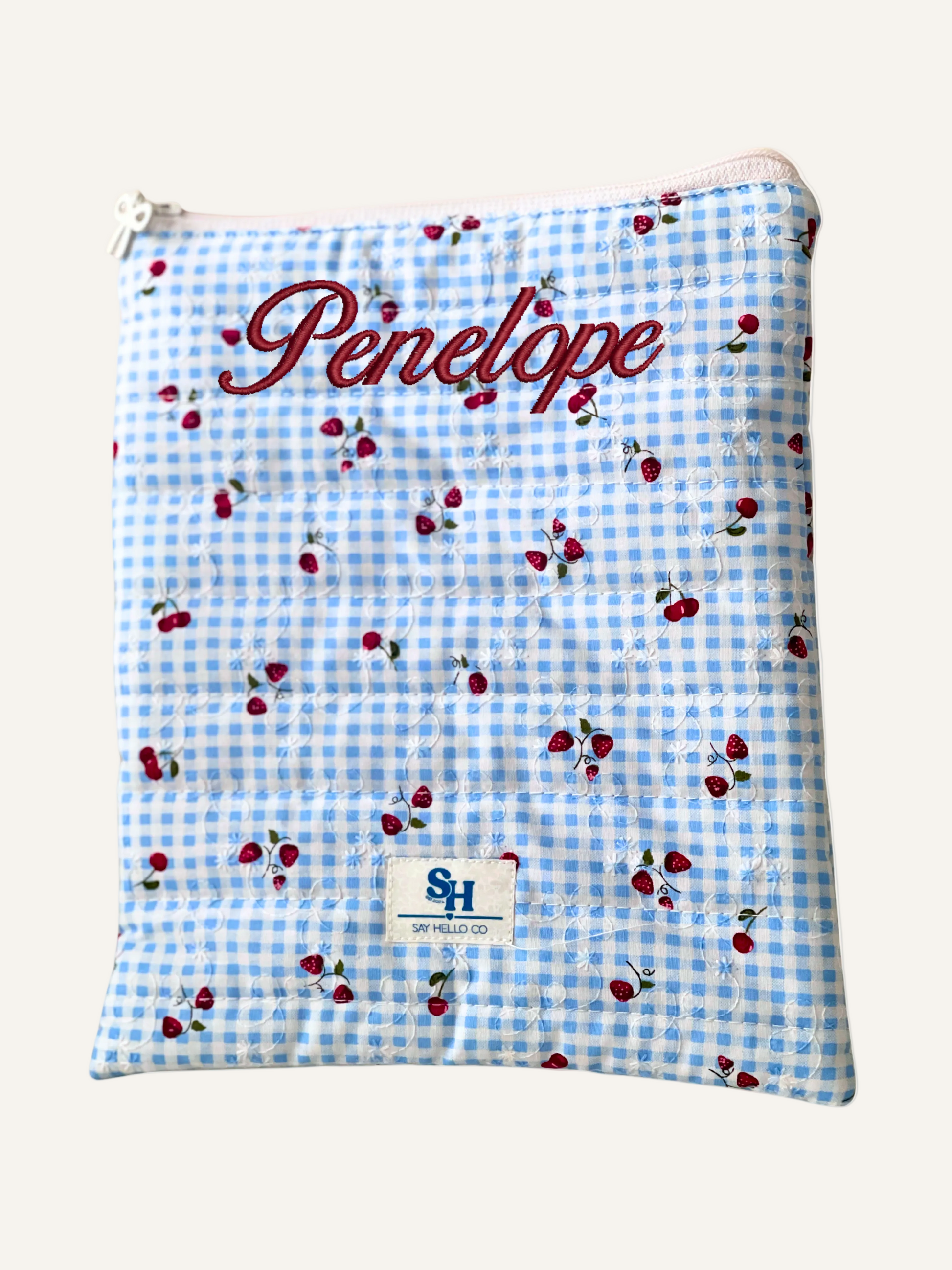 Blue checkered pouch with red cherries and 'Penelope' embroidered cursive text on a warm cream background