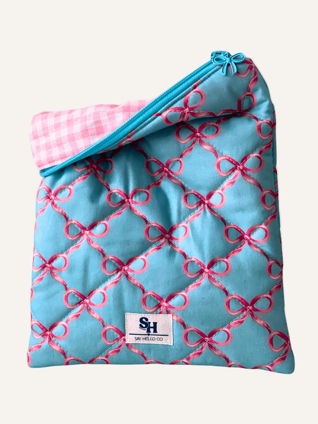 a quilted book sleeve in light blue with pink bows, a blue bow zipper with a pink gingham lining on a warm cream background