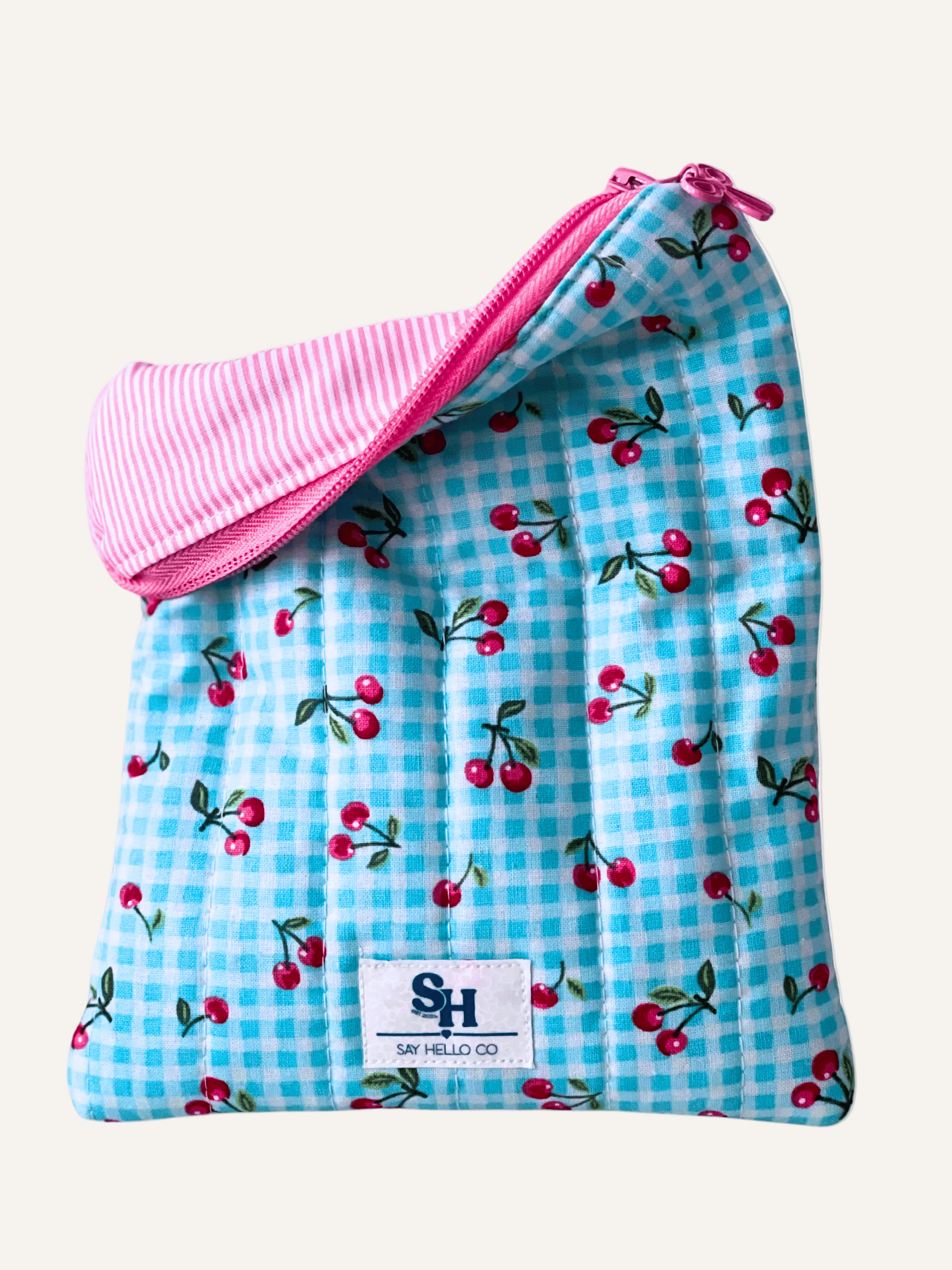 Blue gingham pouch with cherry pattern and pink zipper with a bow zipper pull, pink and cream pinstripe lining featuring a visible brand logo on a warm cream background.