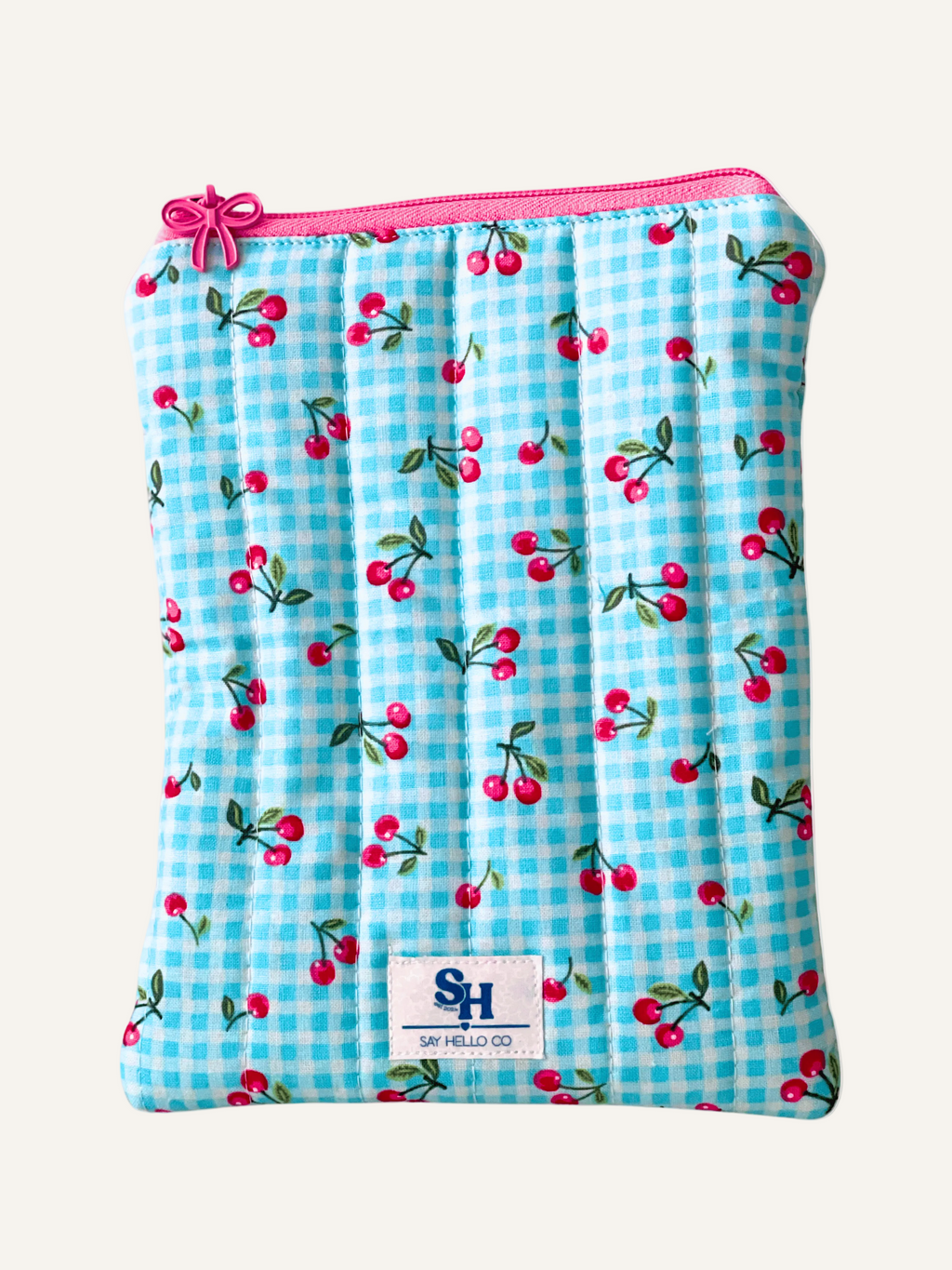 Blue gingham pouch with cherry pattern and pink zipper with a bow zipper pull, featuring a visible brand logo on a warm cream background.