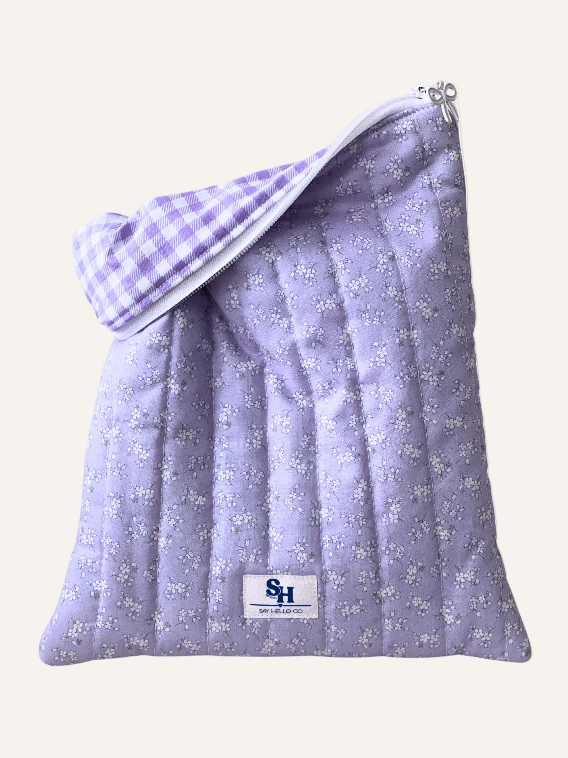 a quilted book sleeve in light lilac with delicate white flowers with light lilac gingham and visible brand label with a white bow zipper on a warm cream background
