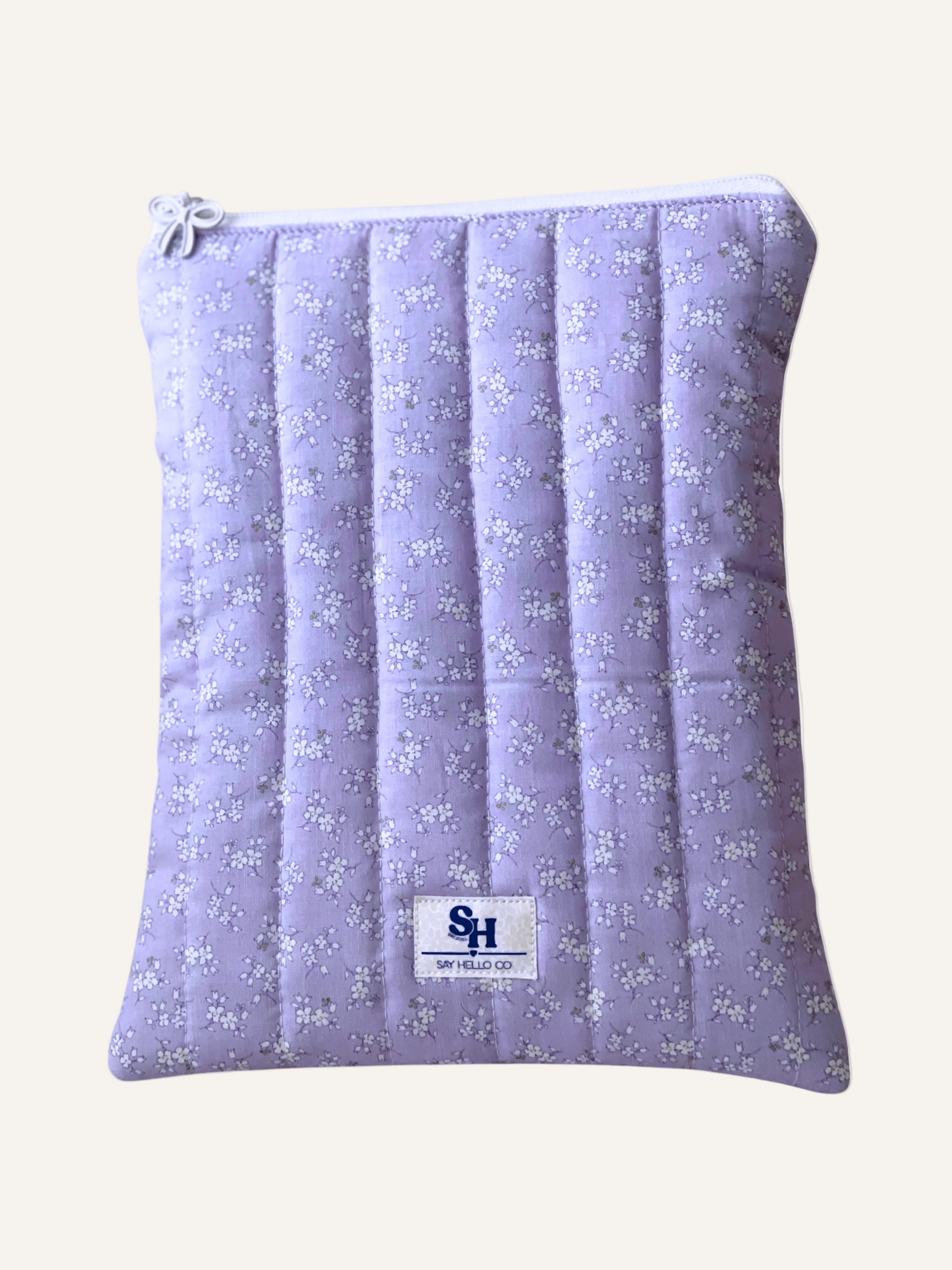 a quilted book sleeve in light lilac with delicate white flowers and visible brand label with a white bow zipper on a warm cream background