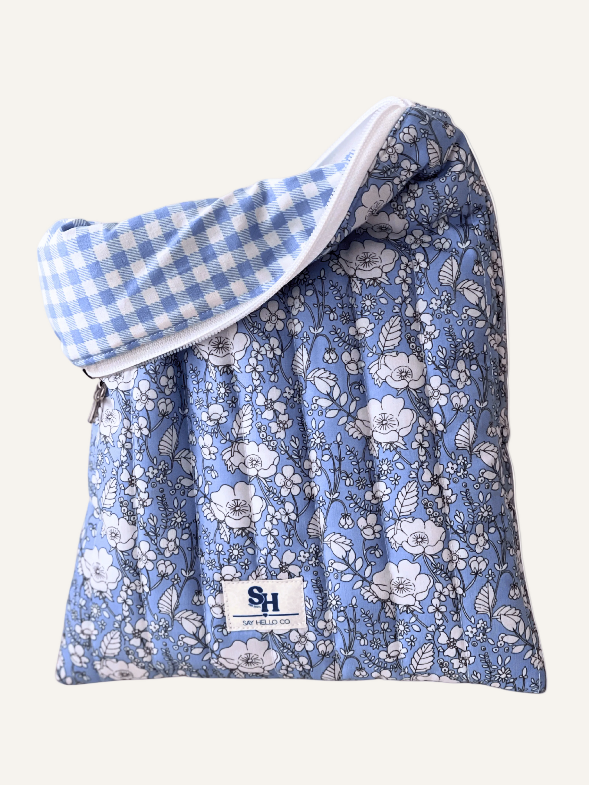 a quilted book sleeve in light blue with white flowers outlined in fine black lines with a silver bow and a visible brand label with a light blue gingham lining on a warm cream background