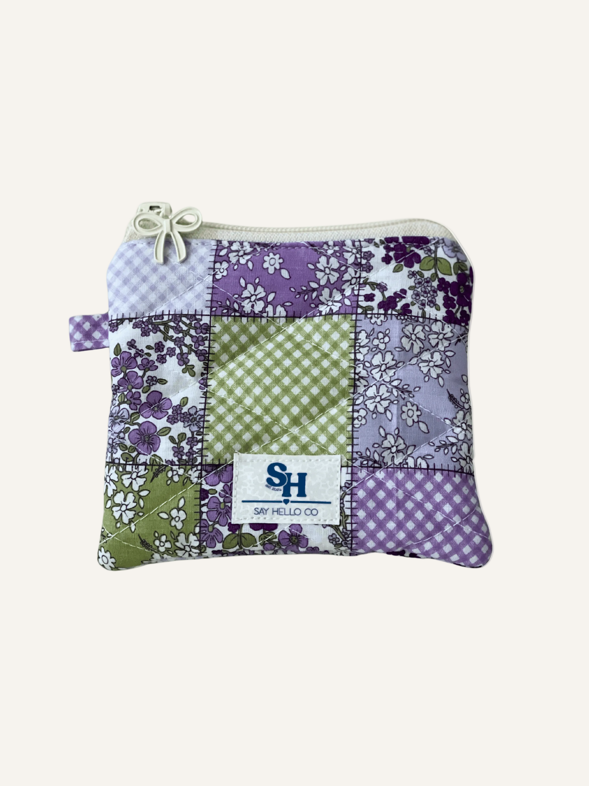 Green, purple and white Patchwork design fabric with floral and gingham patterns as a quilted pouch with a cream bow zipper  on a warm cream background