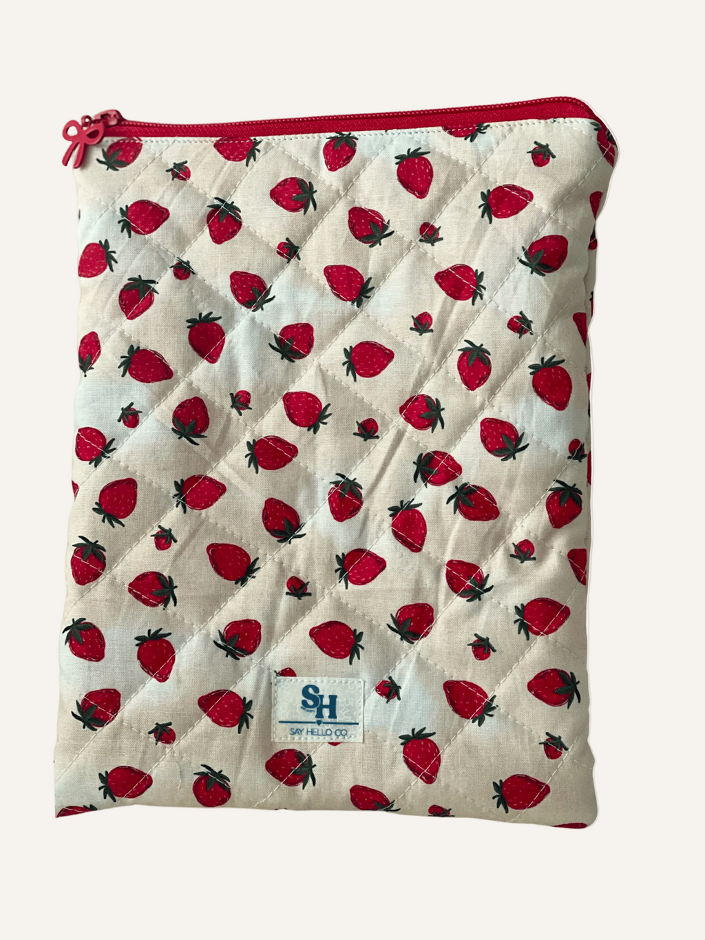 quilted book sleeve in strawberry print on cream with red bow zipper and a visible brand label on a warm cream background