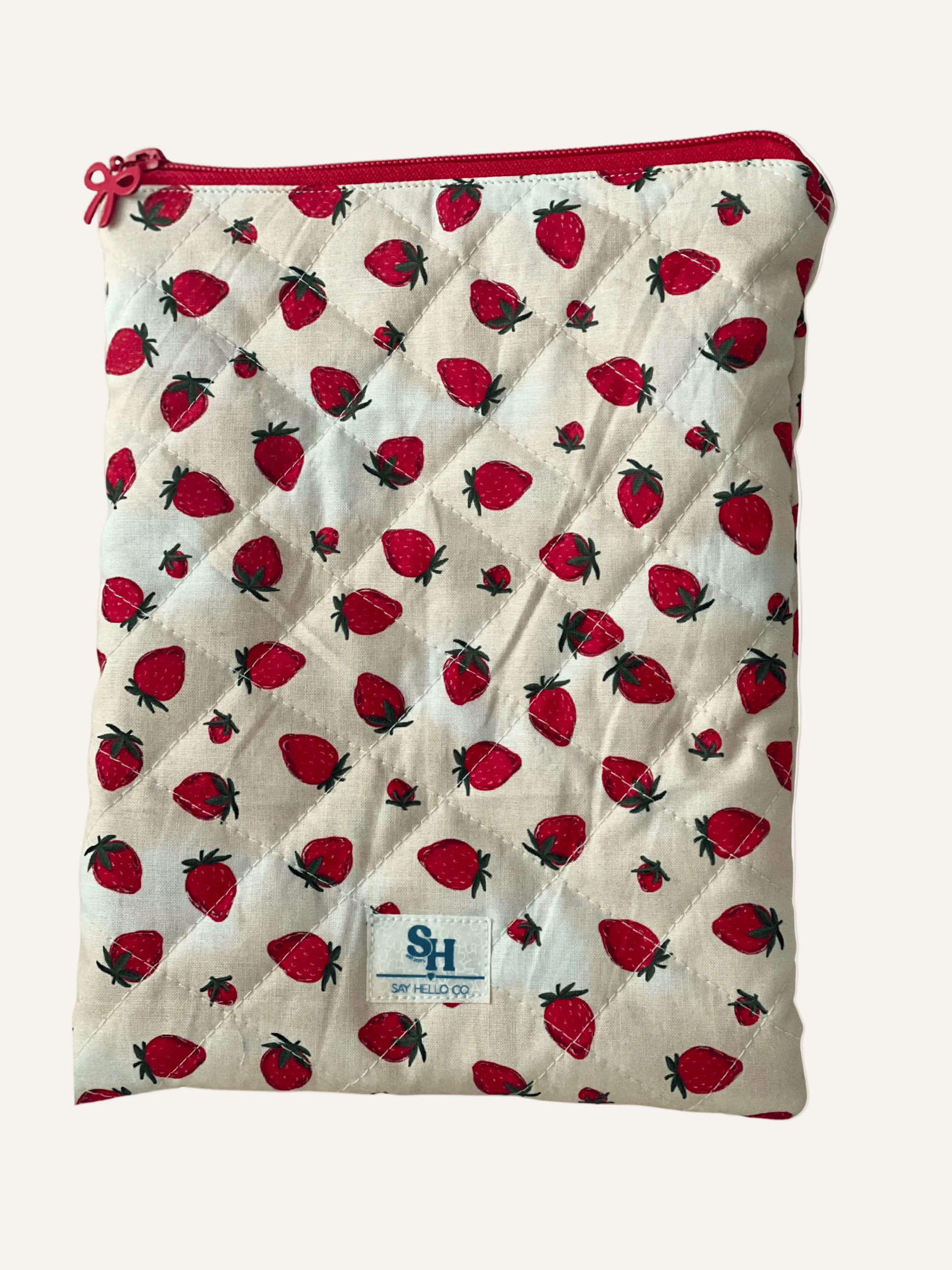 quilted book sleeve in strawberry print on cream with red bow zipper and a visible brand label on a warm cream background