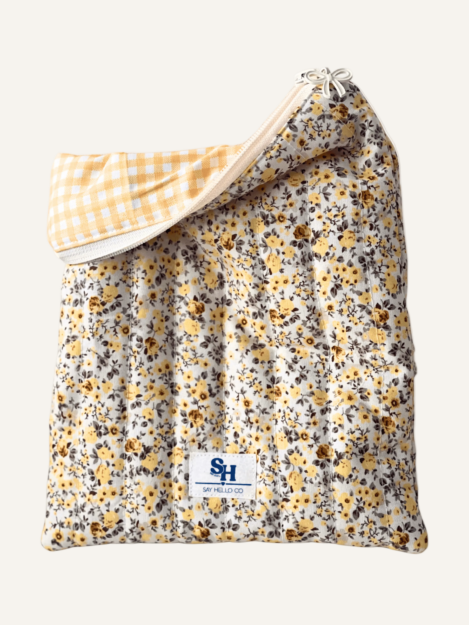 a quilted book sleeve in a ditsy yellow floral print with a cream bow zipper, a visible brand label and yellow gingham lining on a warm cream background