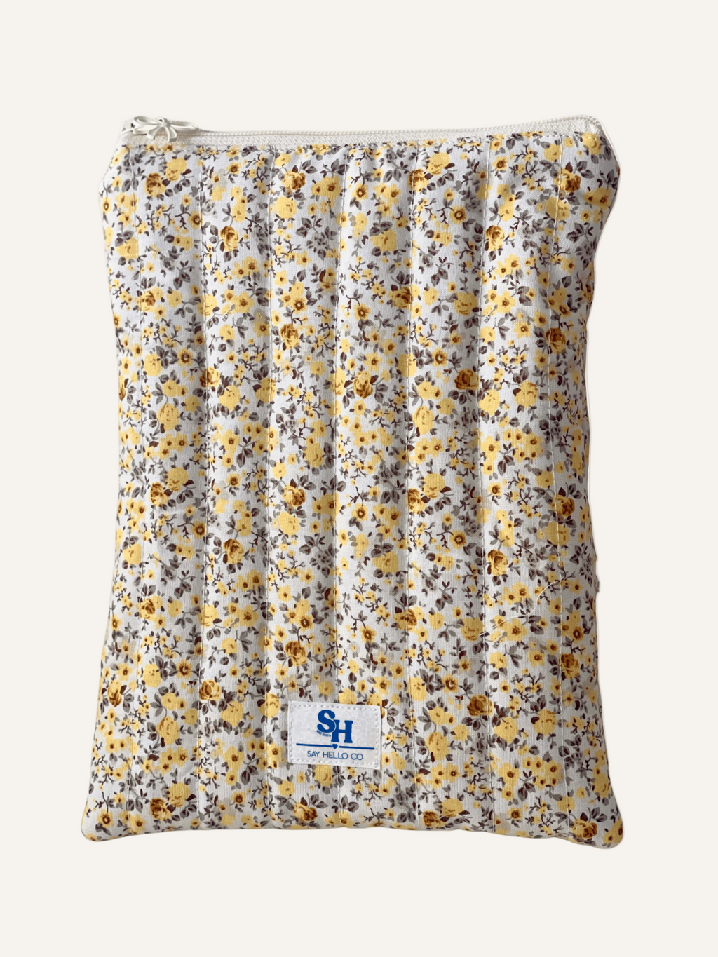 a quilted book sleeve in a ditsy yellow floral print with a cream bow zipper and a visible brand label on a warm cream background