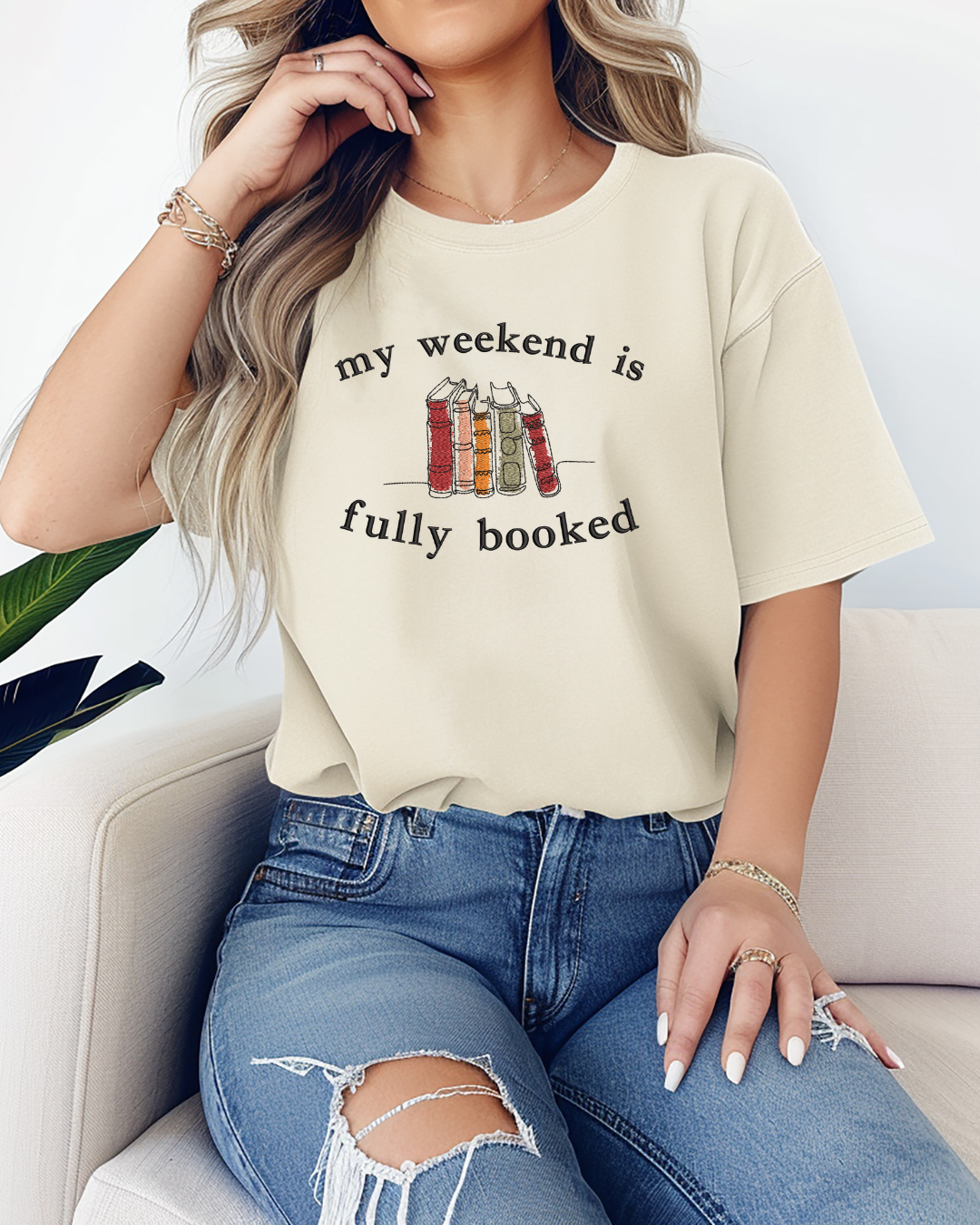 A woman sitting on a couch wearing a beige t-shirt with the phrase 'my weekend is fully booked' embroidered on the front.