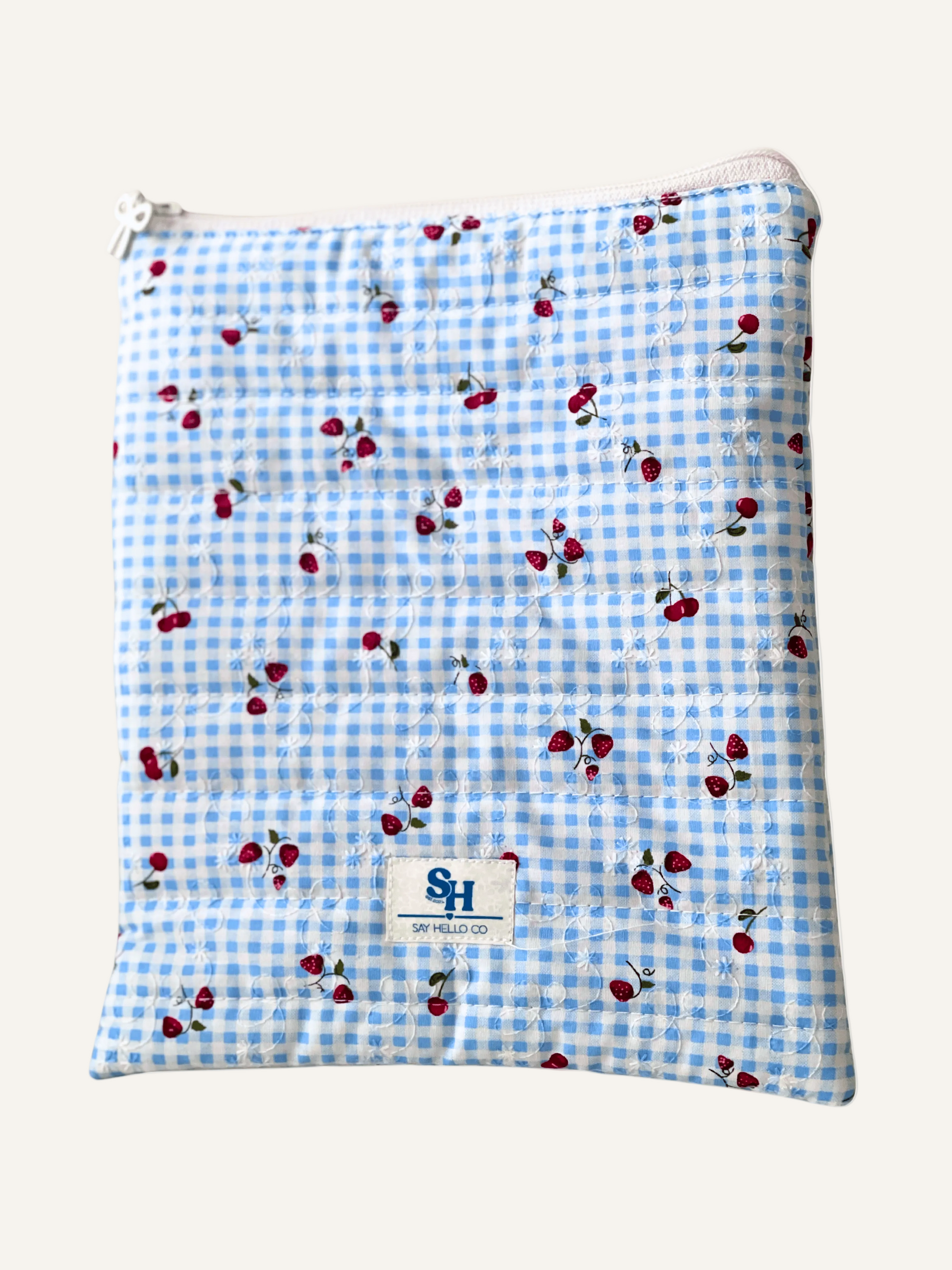 a quilted book sleeve in blue gingham with cherries and strawberries with a white bow zipper and a visible brand label on a warm cream background
