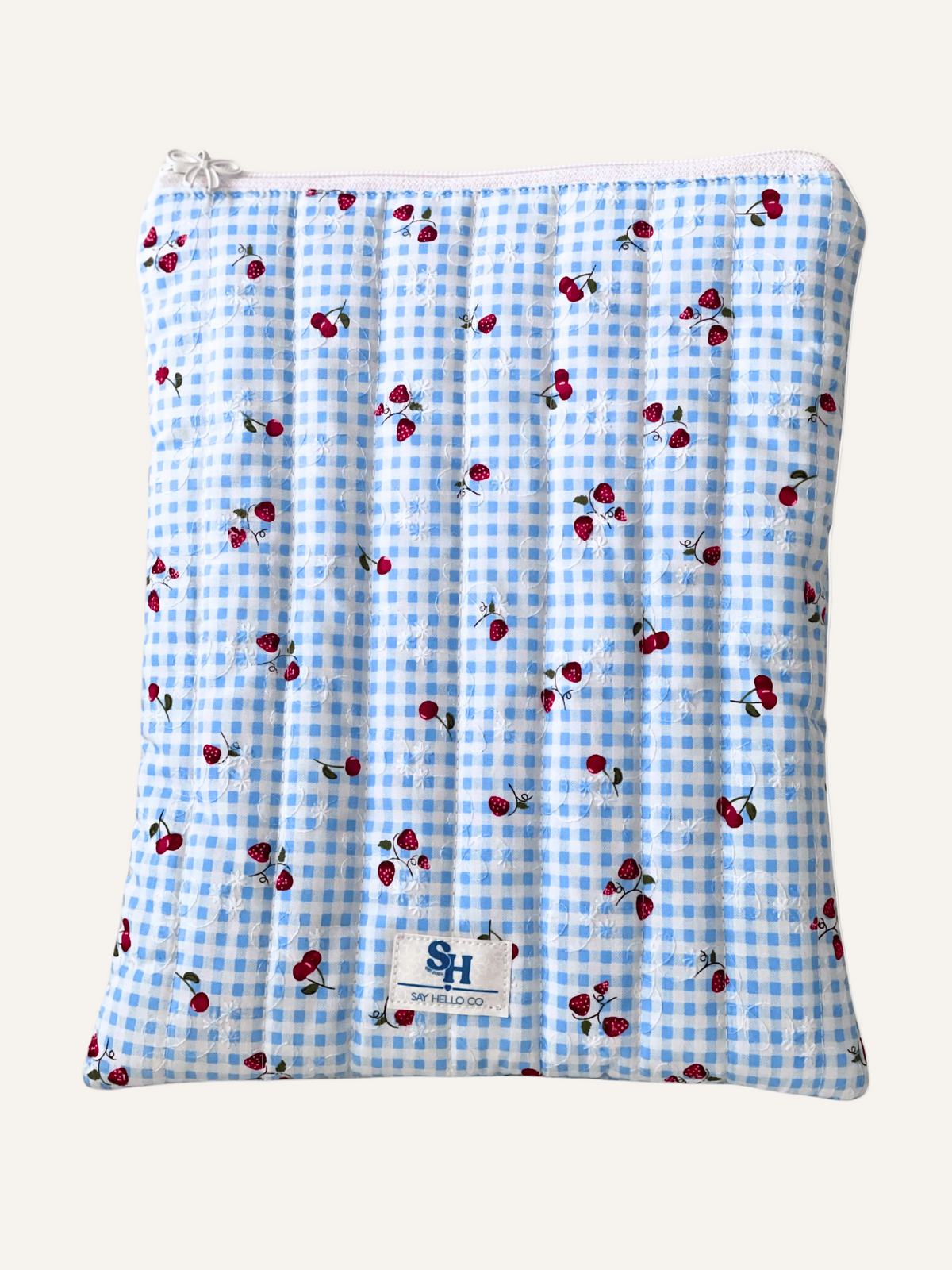 a quilted book sleeve in blue gingham with cherries and strawberries with a white bow zipper and a visible brand label on a warm cream background