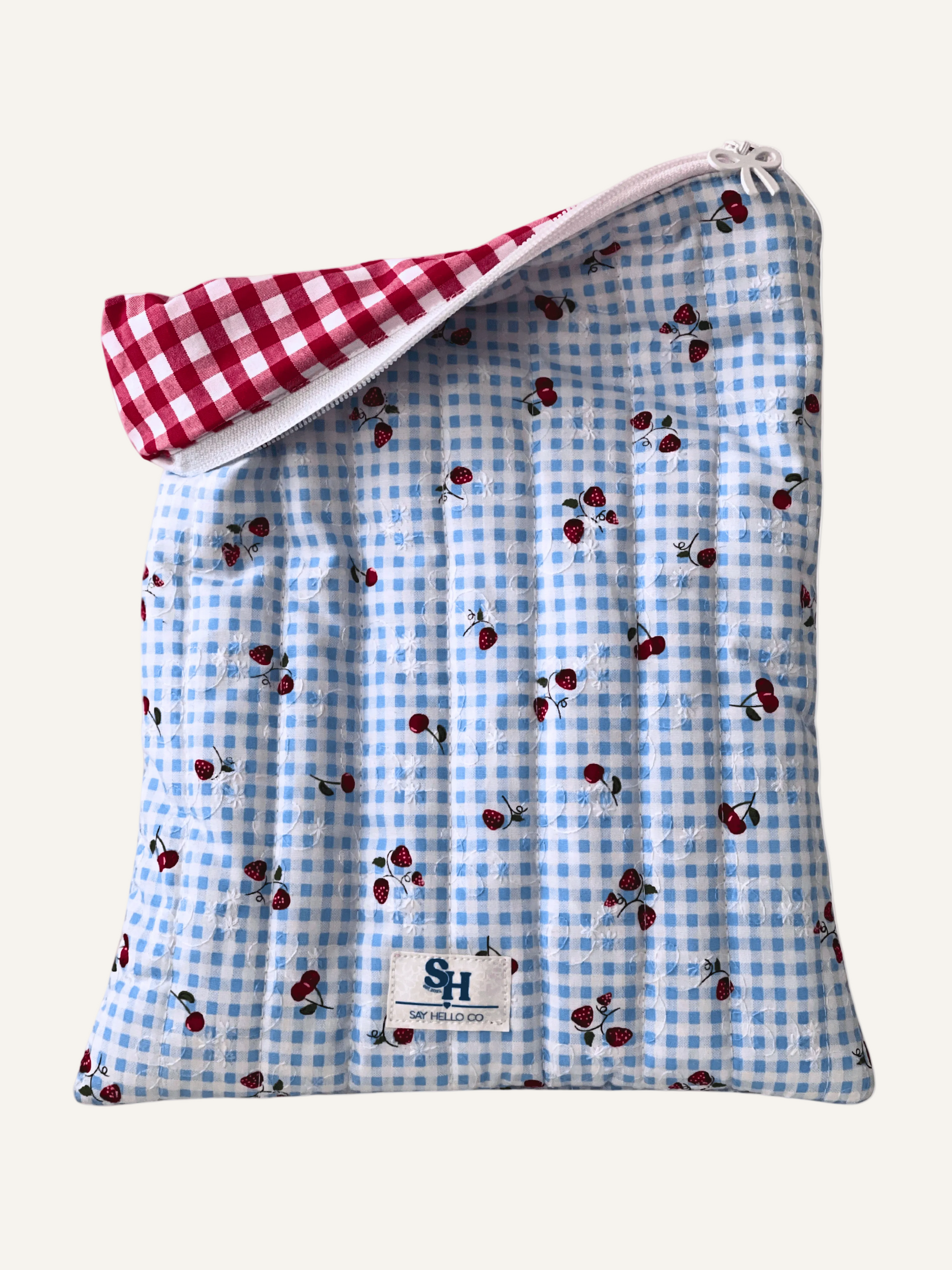 a quilted book sleeve in blue gingham with cherries and strawberries with a white bow zipper, a visible brand label and red gingham lining on a warm cream background