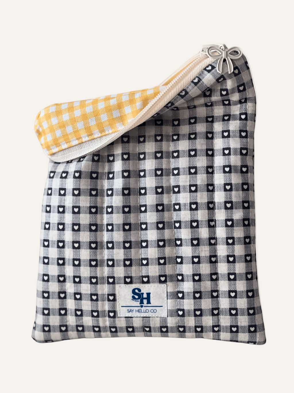 a quilted book sleeve in grey and cream gingham with tiny love hearts, a visible brand label with yellow gingham lining and a cream bow zipper on a warm cream background
