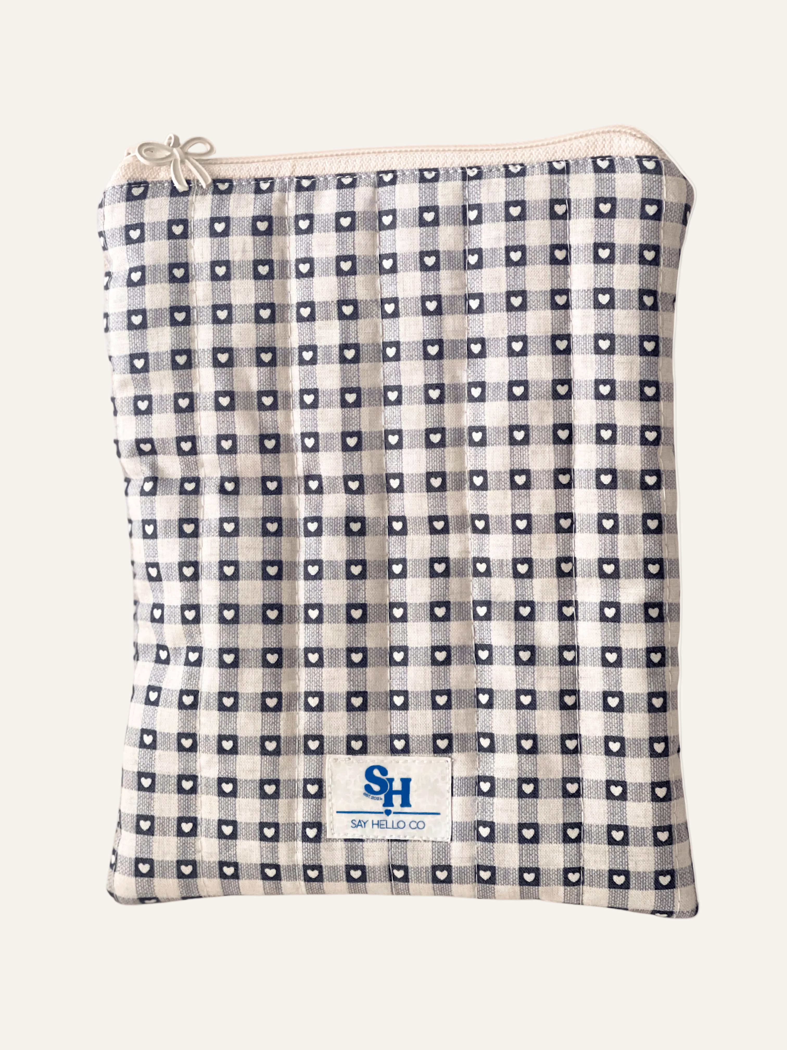 a quilted book sleeve in grey and cream gingham with tiny love hearts, a visible brand label and a cream bow zipper on a warm cream background