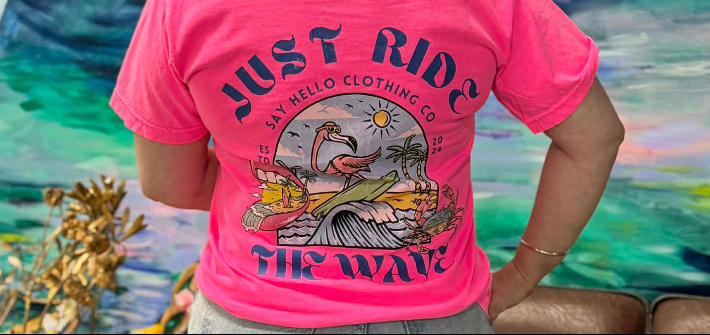Person wearing a pink t-shirt with 'Just Ride the Wave' design in front of a colorful abstract painting.