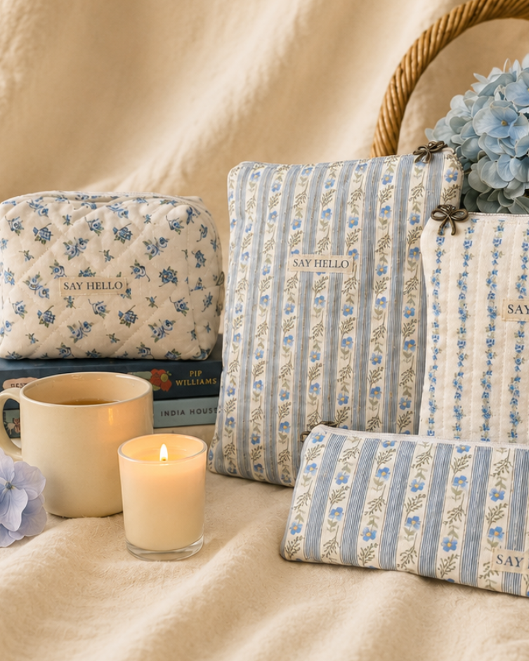sitting on a draped cream linen fabric is some blue and white floral quilted handmade items, book and kindle sleeve, pencil case and a boxy makeup bag sitting on a stack of books, in front of the books is a cup of tea and a lit candle. A hydrangea in a basket is in the background right of the picture. 